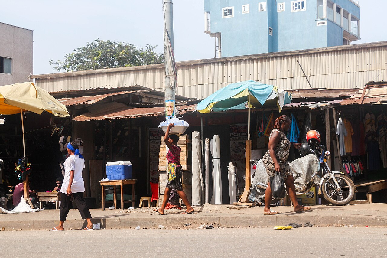 People walking past small businesses on a busy market street in Accra